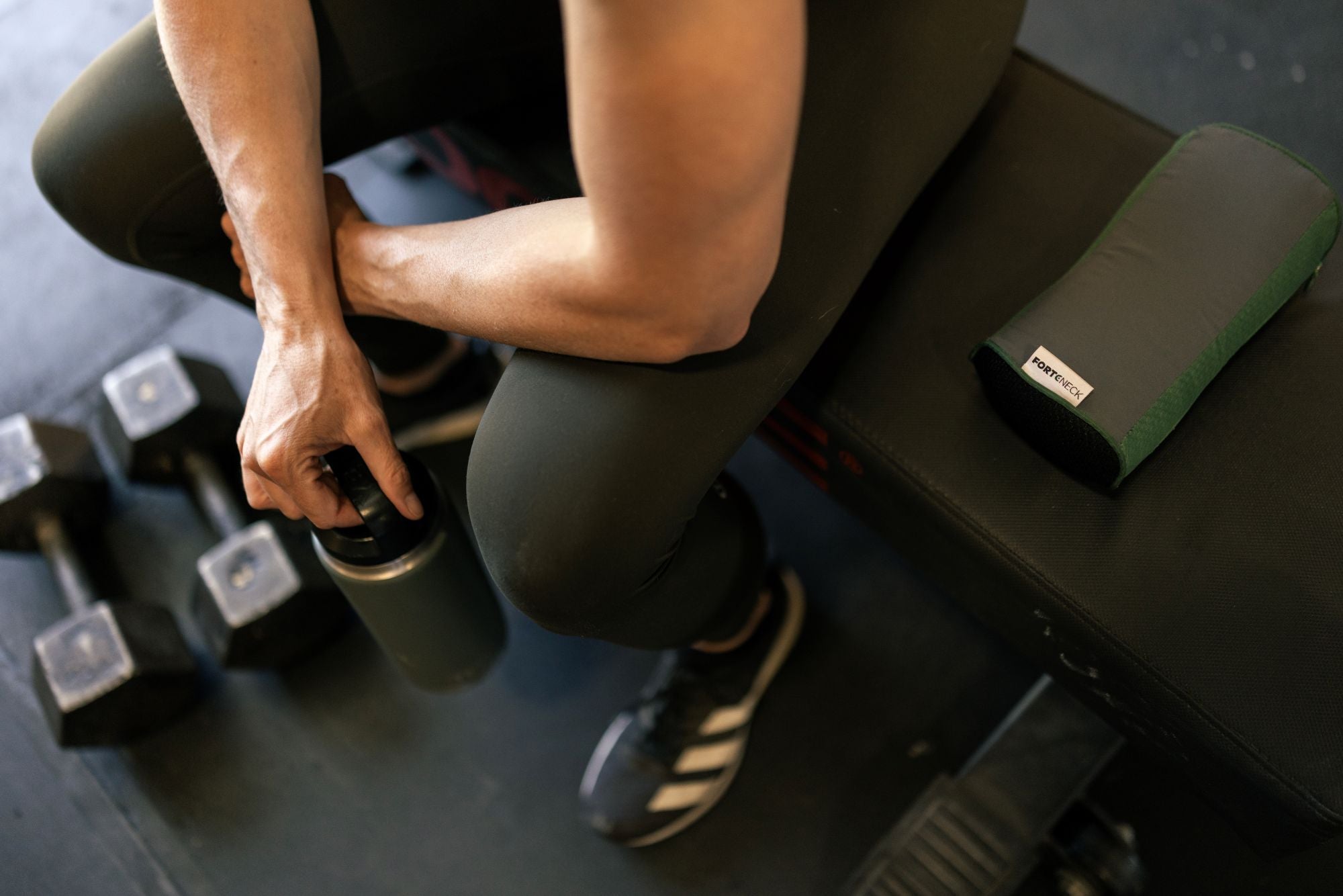 Person sitting on a gym bench with forteneck neck cushion beside her, holding a water bottle and dumbbells on the floor by her feet